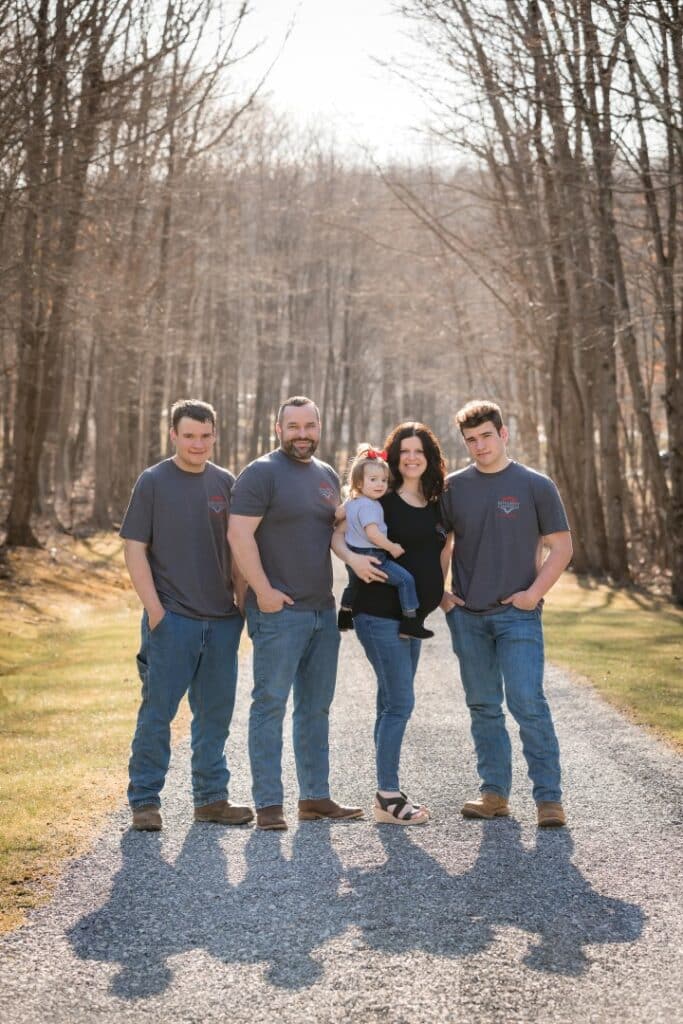 Five people stand on a sunlit path amid leafless trees. Three adults wear matching shirts and jeans; one is holding a young child.