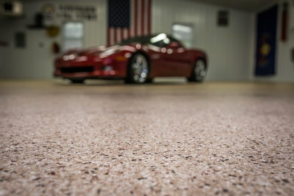 A red sports car is parked in a garage, with the focus on the textured floor in the foreground. Decorative signs and flags adorn the walls.