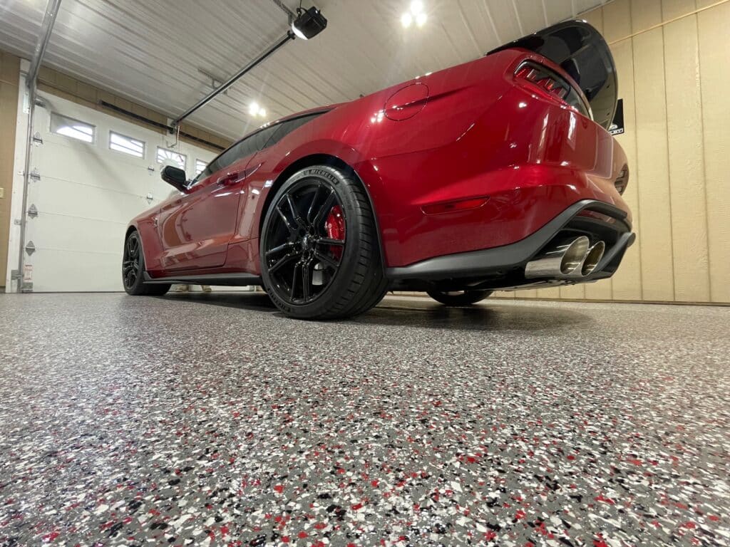 A red sports car with black wheels is parked on a speckled garage floor. It's shown from a rear low-angle perspective, highlighting its sleek design.