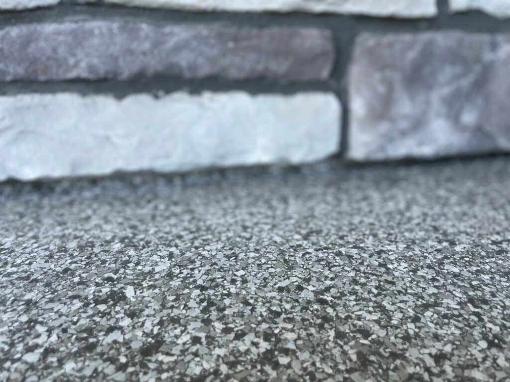 The image shows a close-up of a textured surface meeting a section of a brick wall. The foreground is in focus, emphasizing the granular texture.