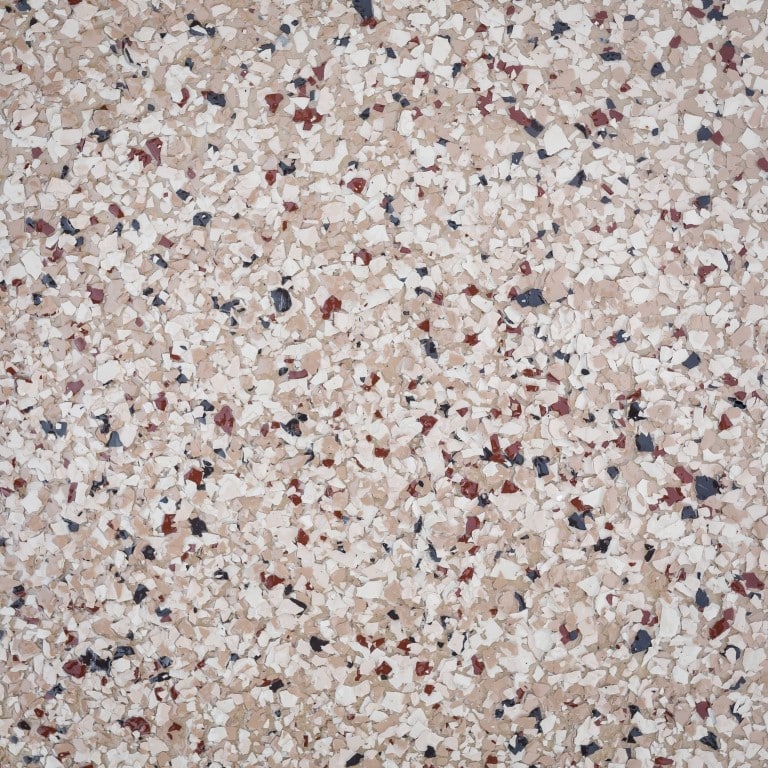 This is a close-up image of a terrazzo floor, comprising numerous small, multicolored chips, predominantly beige, interspersed with red and blue pieces, embedded in concrete.