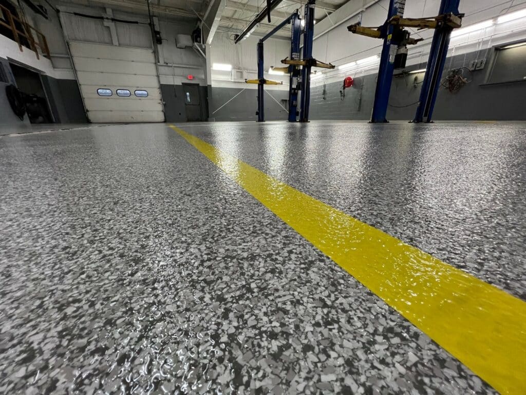 An empty garage with a glossy speckled floor and a bright yellow line. Two blue car lifts are in the background, and garage doors are visible.