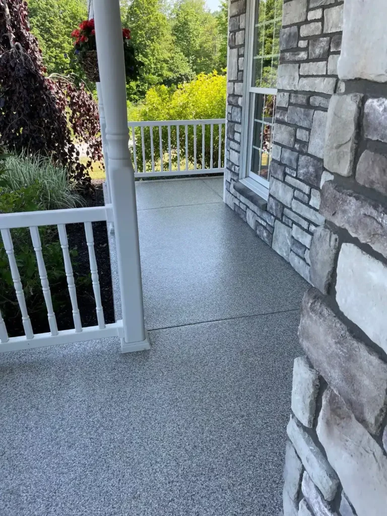 This image shows a corner of a porch with a white railing, gray textured flooring, stone column, hanging plant, and a lush green background.